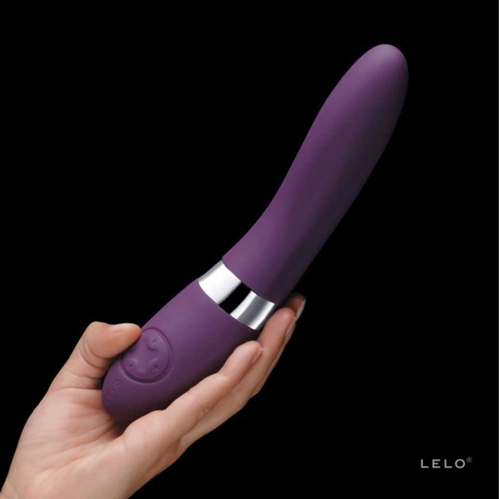 O mână ține un vibrator violet de formă ergonomică, cu detalii argintii, pe un fundal complet negru.